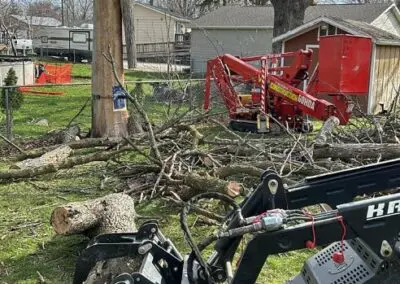 Residential yard with a fallen oak being cut into sections by Vogel Construction & Tree Removal crew using a compact excavator and red stump/chipper machine, scattered logs and branches near a house and chain-link fence in Waterloo, IA