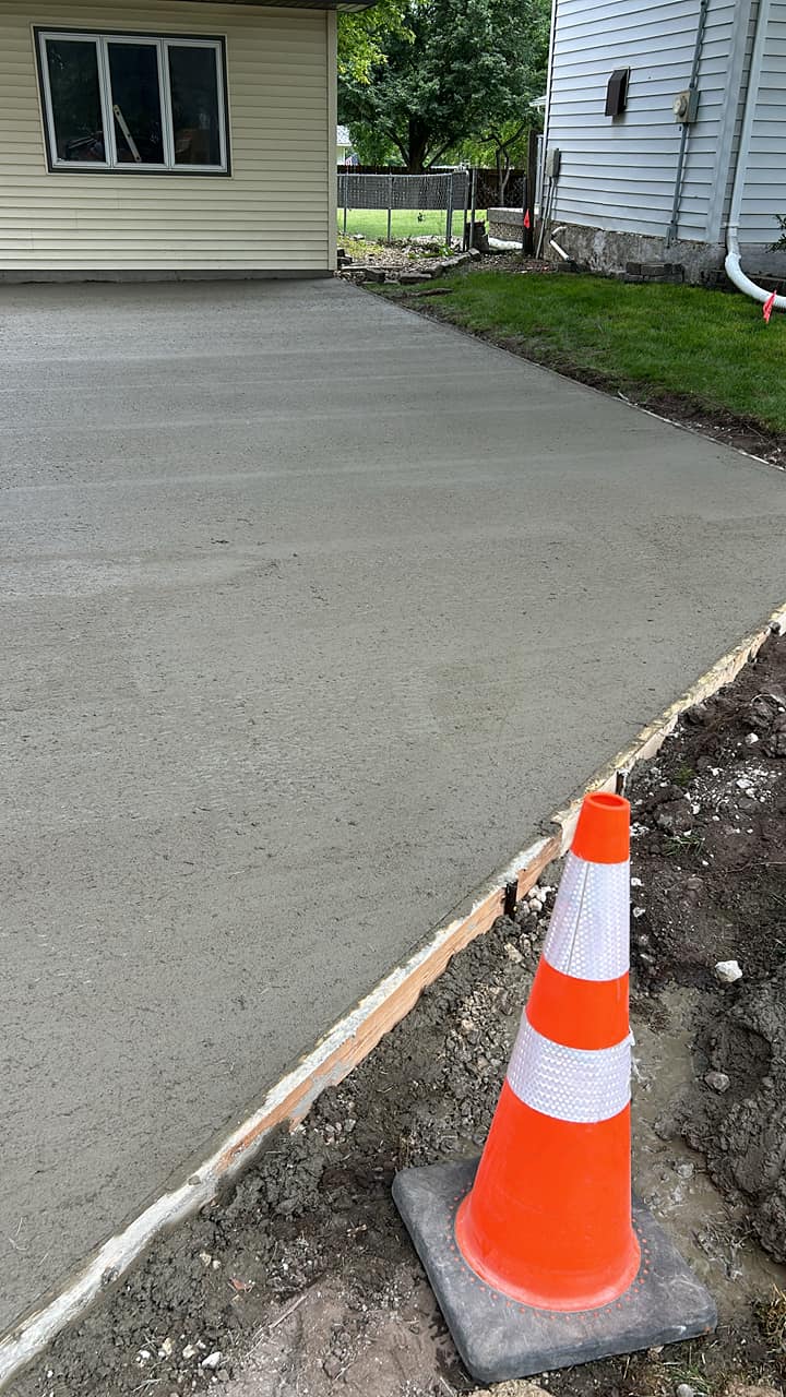 Freshly poured concrete driveway beside two houses with wooden forms and an orange safety cone in the foreground, illustrating Vogel Construction & Tree Removal LLC driveway/excavation work in Waterloo, IA